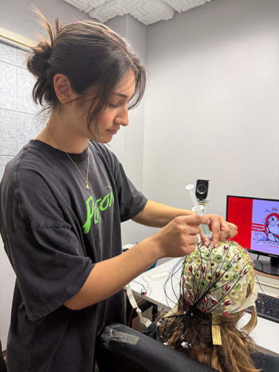 Neuroscience student applies electrodes to EEG cap during sleep research study in a university lab, monitoring brain activity on a nearby computer screen