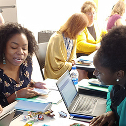 A woman working on a laptop leans over to speak to someone holding a cell phone. Several other people are working in the background.
